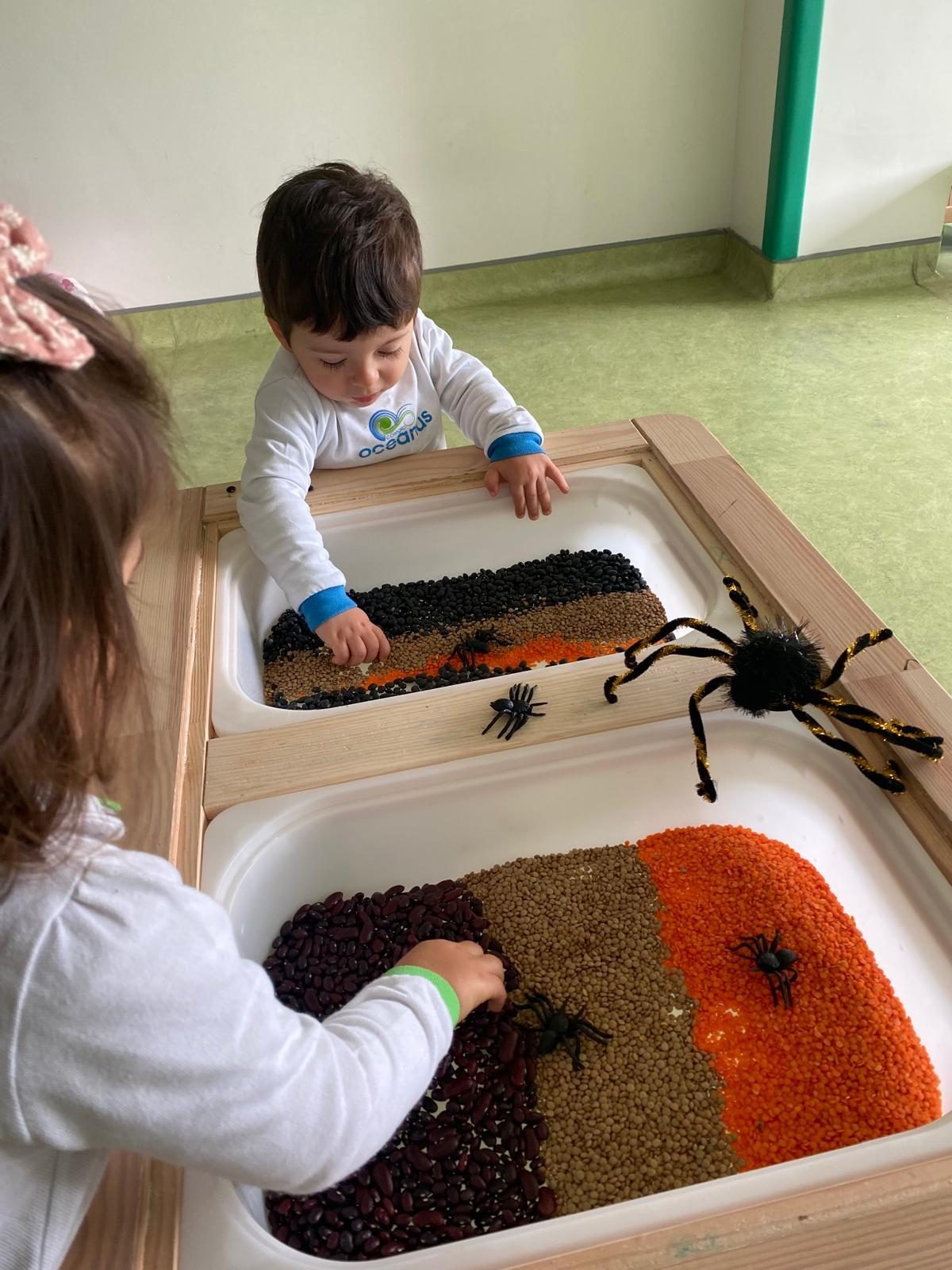 Duas crianças a mexer em feijões e lentilhas, com aranhas de brincar na mesa